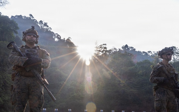Two armed military personnel in full combat gear stand on a grass firing range, with a row of paper shooting targets visible in the background. The scene is backlit by a strong sun flare breaking through trees, creating dramatic lighting with lens flare artifacts. The setting appears to be an outdoor range in a forested, tropical or subtropical environment.