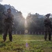 Two armed military personnel in full combat gear stand on a grass firing range, with a row of paper shooting targets visible in the background. The scene is backlit by a strong sun flare breaking through trees, creating dramatic lighting with lens flare artifacts. The setting appears to be an outdoor range in a forested, tropical or subtropical environment.