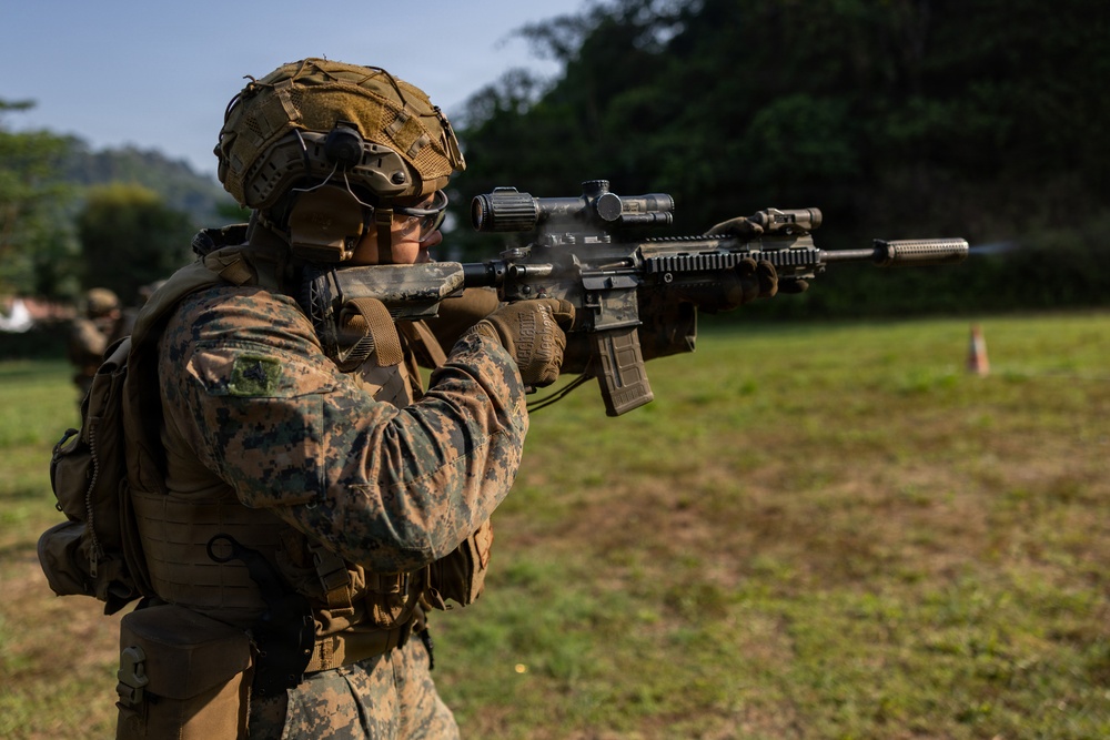 A military service member in MARPAT (Marine pattern) digital camouflage uniform is actively firing a rifle at an outdoor shooting range. The weapon appears to be an M4/M16-style carbine equipped with an optic scope and tactical accessories, with visible muzzle smoke indicating recent discharge. A second service member is partially visible in the background, suggesting a group training exercise.