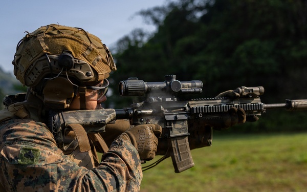 A military service member in MARPAT (Marine pattern) digital camouflage uniform is actively firing a rifle at an outdoor shooting range. The weapon appears to be an M4/M16-style carbine equipped with an optic scope and tactical accessories, with visible muzzle smoke indicating recent discharge. A second service member is partially visible in the background, suggesting a group training exercise.