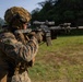 A military service member in MARPAT (Marine pattern) digital camouflage uniform is actively firing a rifle at an outdoor shooting range. The weapon appears to be an M4/M16-style carbine equipped with an optic scope and tactical accessories, with visible muzzle smoke indicating recent discharge. A second service member is partially visible in the background, suggesting a group training exercise.