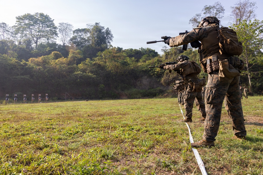 A line of military personnel in full combat gear are conducting a live-fire range exercise on an outdoor shooting range. They are advancing in a column formation along a marked lane, aiming rifles at paper silhouette targets positioned downrange. The setting appears to be a tropical or subtropical environment based on the surrounding vegetation.