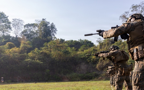 A line of military personnel in full combat gear are conducting a live-fire range exercise on an outdoor shooting range. They are advancing in a column formation along a marked lane, aiming rifles at paper silhouette targets positioned downrange. The setting appears to be a tropical or subtropical environment based on the surrounding vegetation.