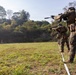 A line of military personnel in full combat gear are conducting a live-fire range exercise on an outdoor shooting range. They are advancing in a column formation along a marked lane, aiming rifles at paper silhouette targets positioned downrange. The setting appears to be a tropical or subtropical environment based on the surrounding vegetation.