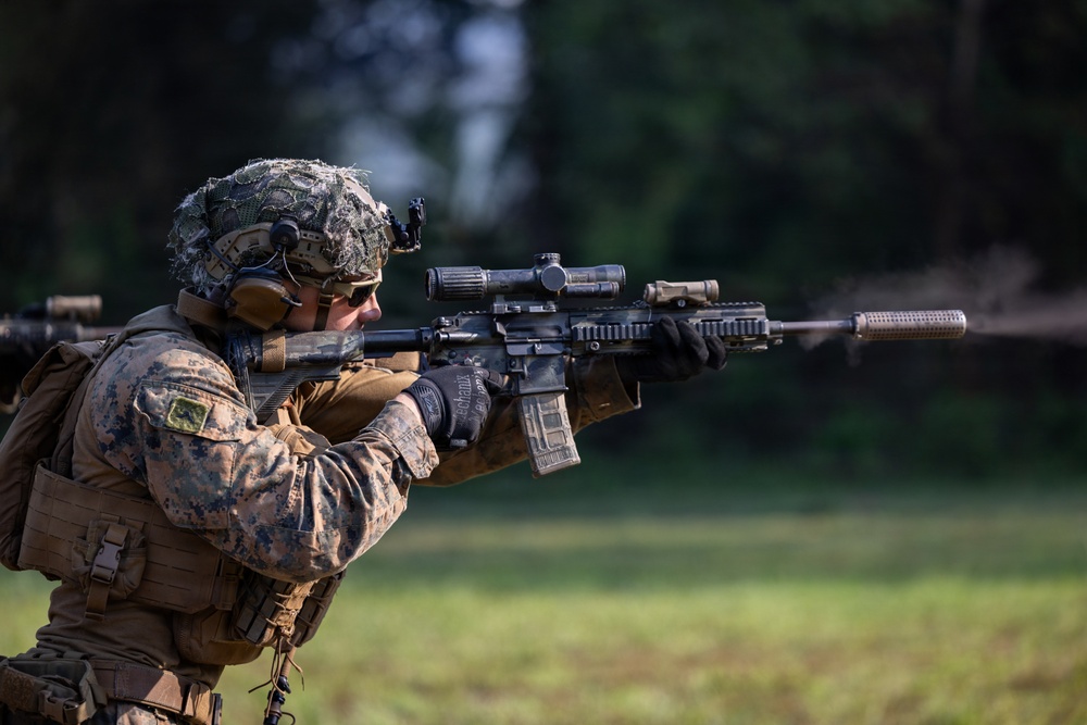 A U.S. Marine in full combat gear is actively firing a suppressed rifle at a shooting range or training area. The soldier is wearing MARPAT (Marine Pattern) camouflage utilities, a camouflage-netted helmet, and tactical equipment. Visible muzzle blast distortion can be seen along the barrel, indicating the weapon has just been discharged.