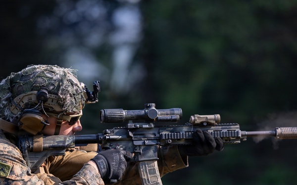A U.S. Marine in full combat gear is actively firing a suppressed rifle at a shooting range or training area. The soldier is wearing MARPAT (Marine Pattern) camouflage utilities, a camouflage-netted helmet, and tactical equipment. Visible muzzle blast distortion can be seen along the barrel, indicating the weapon has just been discharged.