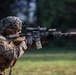 A U.S. Marine in full combat gear is actively firing a suppressed rifle at a shooting range or training area. The soldier is wearing MARPAT (Marine Pattern) camouflage utilities, a camouflage-netted helmet, and tactical equipment. Visible muzzle blast distortion can be seen along the barrel, indicating the weapon has just been discharged.