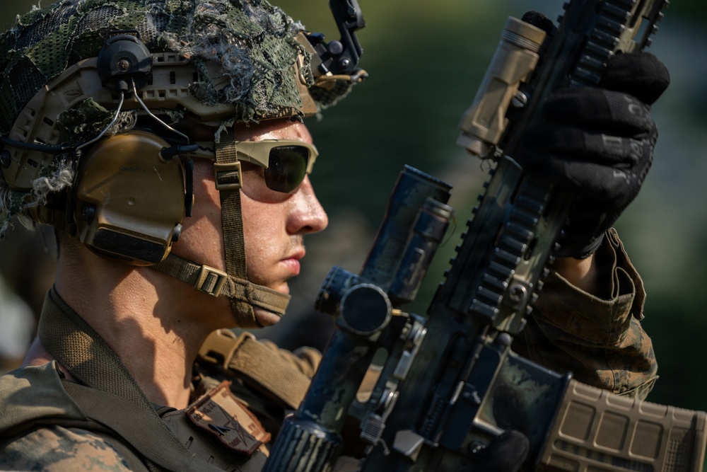 A close-up photograph of a military service member in full combat gear examining or inspecting a rifle. The subject is wearing MARPAT (Marine pattern) camouflage uniform and helmet with netting, tactical hearing protection, and ballistic sunglasses. The image is professionally composed with sharp focus on the subject against a blurred green background.