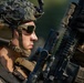 A close-up photograph of a military service member in full combat gear examining or inspecting a rifle. The subject is wearing MARPAT (Marine pattern) camouflage uniform and helmet with netting, tactical hearing protection, and ballistic sunglasses. The image is professionally composed with sharp focus on the subject against a blurred green background.