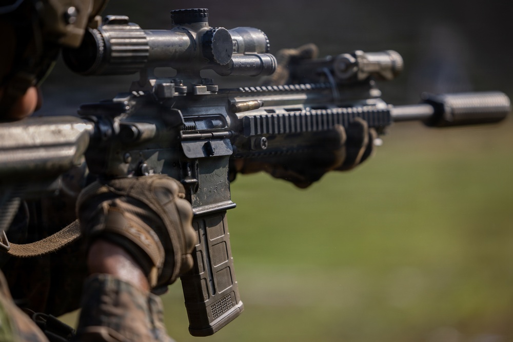 A close-up photograph of a military or law enforcement operator holding an AR-platform rifle with multiple accessories mounted. The shooter is wearing camouflage uniform and tactical gloves, aiming the weapon downrange. The image is a sharp, professional-quality photograph focused on the weapon system.