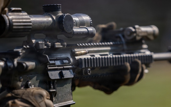 A close-up photograph of a military or law enforcement operator holding an AR-platform rifle with multiple accessories mounted. The shooter is wearing camouflage uniform and tactical gloves, aiming the weapon downrange. The image is a sharp, professional-quality photograph focused on the weapon system.