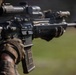 A close-up photograph of a military or law enforcement operator holding an AR-platform rifle with multiple accessories mounted. The shooter is wearing camouflage uniform and tactical gloves, aiming the weapon downrange. The image is a sharp, professional-quality photograph focused on the weapon system.