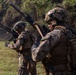 Two military personnel in full combat gear are positioned in an open grassy field with dense vegetation in the background. Both are wearing MARPAT (Marine Pattern) camouflage uniforms, tactical helmets, body armor, and carrying rifles, consistent with U.S. Marine Corps equipment. The foreground soldier has a name tape reading 'CASTILLO' visible on their backpack.