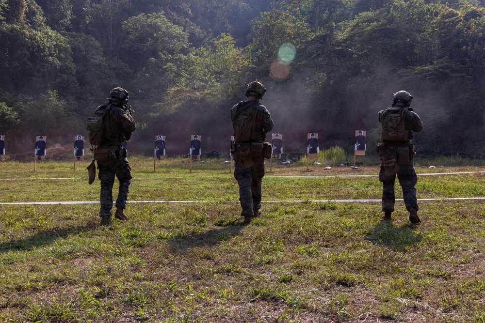 Three military personnel in full combat gear stand at a firing range, facing a row of silhouette paper targets downrange. The scene appears to be an outdoor marksmanship training exercise, with smoke or dust visible in the background near the target line. The setting is a wooded, open-field range with numbered target stands.