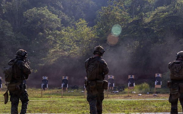 Three military personnel in full combat gear stand at a firing range, facing a row of silhouette paper targets downrange. The scene appears to be an outdoor marksmanship training exercise, with smoke or dust visible in the background near the target line. The setting is a wooded, open-field range with numbered target stands.