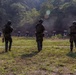 Three military personnel in full combat gear stand at a firing range, facing a row of silhouette paper targets downrange. The scene appears to be an outdoor marksmanship training exercise, with smoke or dust visible in the background near the target line. The setting is a wooded, open-field range with numbered target stands.