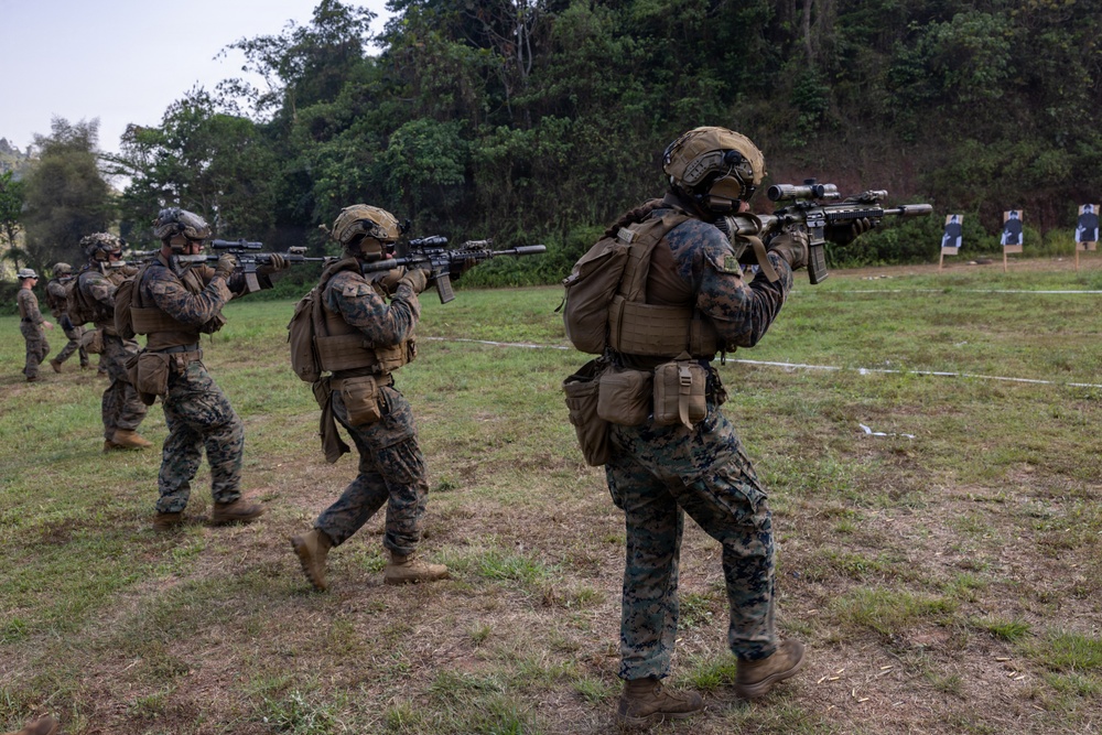 A group of military personnel in full combat gear are conducting a live-fire range exercise on an outdoor grass field. The soldiers, wearing MARPAT (Marine pattern) camouflage uniforms, are advancing in a line formation while aiming rifles at paper targets positioned to the right. The setting appears to be a training range surrounded by dense tropical or subtropical vegetation.