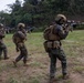 A group of military personnel in full combat gear are conducting a live-fire range exercise on an outdoor grass field. The soldiers, wearing MARPAT (Marine pattern) camouflage uniforms, are advancing in a line formation while aiming rifles at paper targets positioned to the right. The setting appears to be a training range surrounded by dense tropical or subtropical vegetation.