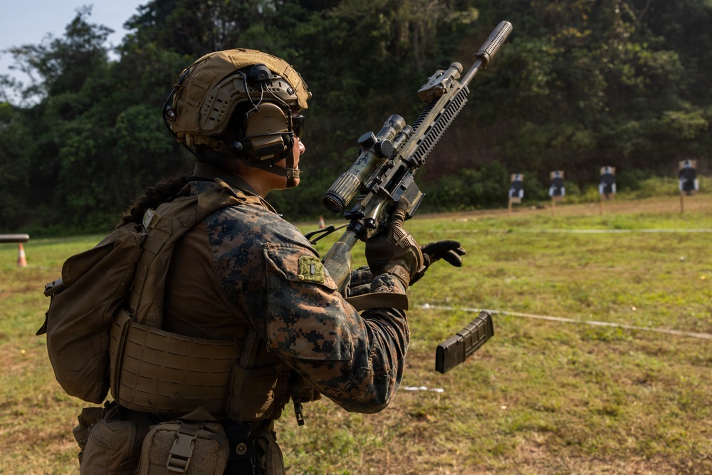 A military service member in full combat gear is conducting a weapons reload or magazine change drill at an outdoor shooting range. The individual is wearing MARPAT (Marine Pattern) camouflage, indicating affiliation with the United States Marine Corps. Silhouette targets are visible downrange in the background.
