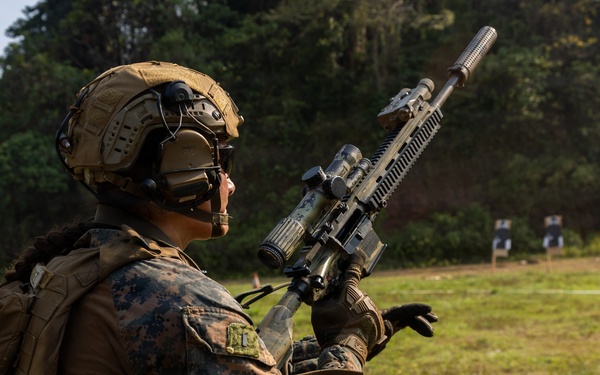 A military service member in full combat gear is conducting a weapons reload or magazine change drill at an outdoor shooting range. The individual is wearing MARPAT (Marine Pattern) camouflage, indicating affiliation with the United States Marine Corps. Silhouette targets are visible downrange in the background.
