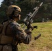 A military service member in full combat gear is conducting a weapons reload or magazine change drill at an outdoor shooting range. The individual is wearing MARPAT (Marine Pattern) camouflage, indicating affiliation with the United States Marine Corps. Silhouette targets are visible downrange in the background.