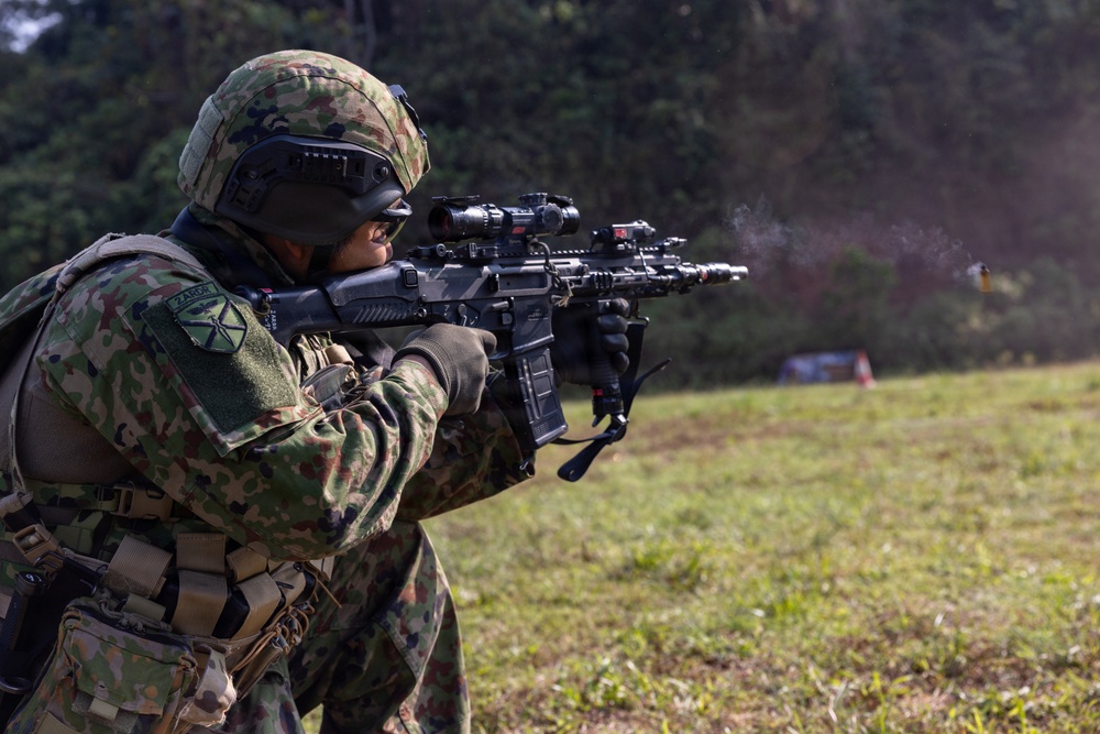 A soldier in camouflage uniform is actively firing a rifle on an outdoor range or field. A spent brass casing is visibly ejecting from the weapon, and a smoke plume is visible near the muzzle. The soldier is equipped with full tactical gear including helmet, optics, and body armor.