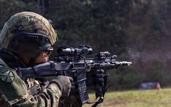 A soldier in camouflage uniform is actively firing a rifle on an outdoor range or field. A spent brass casing is visibly ejecting from the weapon, and a smoke plume is visible near the muzzle. The soldier is equipped with full tactical gear including helmet, optics, and body armor.