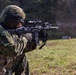A soldier in camouflage uniform is actively firing a rifle on an outdoor range or field. A spent brass casing is visibly ejecting from the weapon, and a smoke plume is visible near the muzzle. The soldier is equipped with full tactical gear including helmet, optics, and body armor.