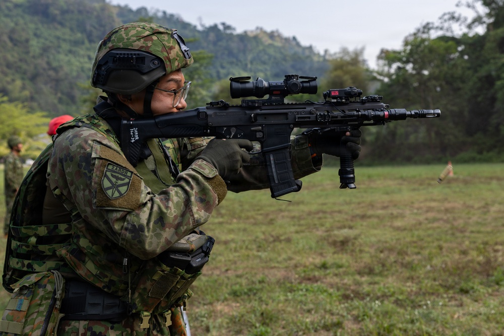 A soldier in camouflage uniform is aiming a modern assault rifle equipped with multiple optics and accessories at a firing range. The soldier wears a tactical helmet with goggles, glasses, and a patch reading '3ARDB' on the sleeve. The setting appears to be an outdoor military training exercise in a green, hilly environment.