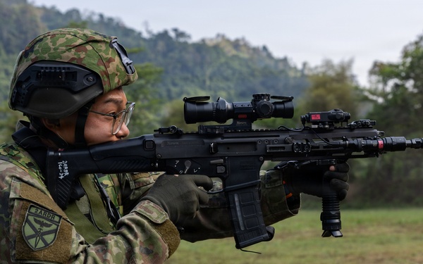 A soldier in camouflage uniform is aiming a modern assault rifle equipped with multiple optics and accessories at a firing range. The soldier wears a tactical helmet with goggles, glasses, and a patch reading '3ARDB' on the sleeve. The setting appears to be an outdoor military training exercise in a green, hilly environment.