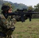 A soldier in camouflage uniform is aiming a modern assault rifle equipped with multiple optics and accessories at a firing range. The soldier wears a tactical helmet with goggles, glasses, and a patch reading '3ARDB' on the sleeve. The setting appears to be an outdoor military training exercise in a green, hilly environment.