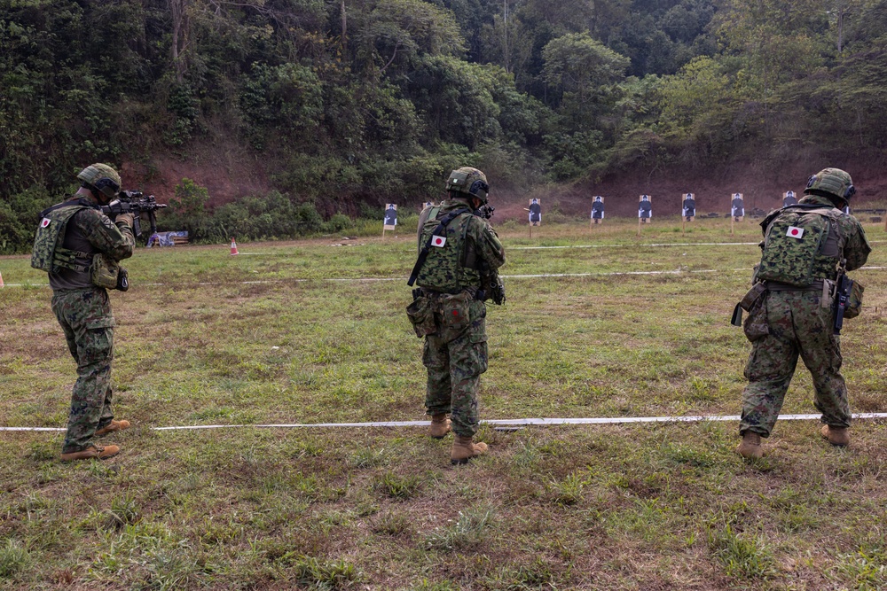 Three military personnel wearing Japanese Ground Self-Defense Force uniforms and tactical gear are positioned at an outdoor shooting range. One soldier on the left is actively aiming a rifle downrange at humanoid paper targets. The setting appears to be a live-fire training exercise at a range bordered by a forested hillside.