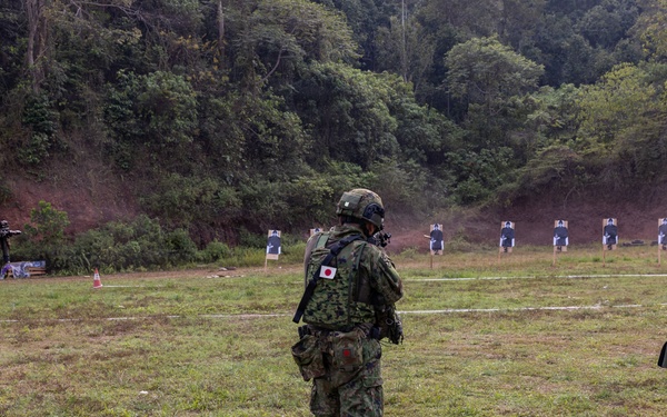 Three military personnel wearing Japanese Ground Self-Defense Force uniforms and tactical gear are positioned at an outdoor shooting range. One soldier on the left is actively aiming a rifle downrange at humanoid paper targets. The setting appears to be a live-fire training exercise at a range bordered by a forested hillside.