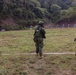 Three military personnel wearing Japanese Ground Self-Defense Force uniforms and tactical gear are positioned at an outdoor shooting range. One soldier on the left is actively aiming a rifle downrange at humanoid paper targets. The setting appears to be a live-fire training exercise at a range bordered by a forested hillside.
