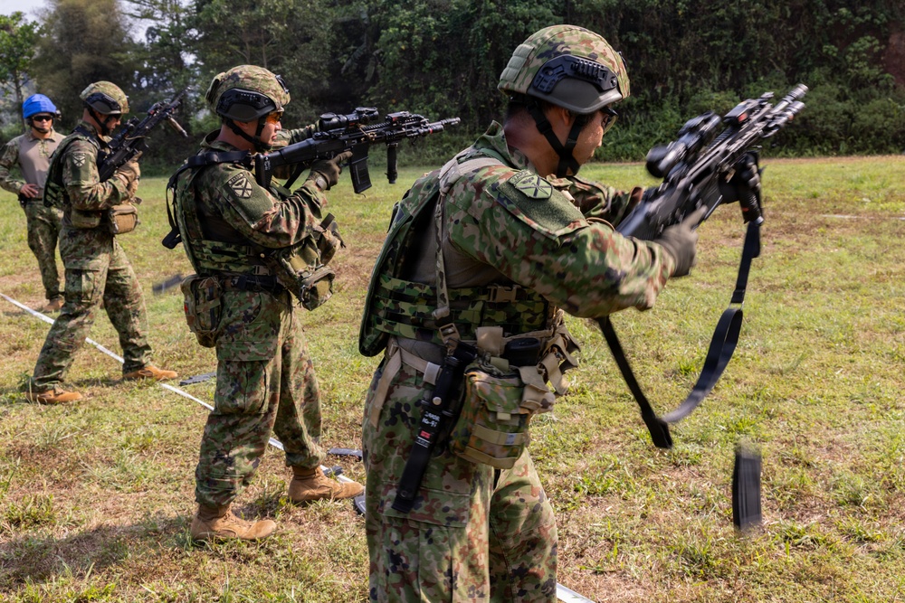 Military personnel in camouflage uniforms are conducting what appears to be a weapons training or demonstration exercise in an outdoor field setting. The soldiers are wearing tactical gear including helmets and body armor, and are handling rifles in various positions. A mix of personnel with different uniform patterns suggests a multinational or joint training exercise.