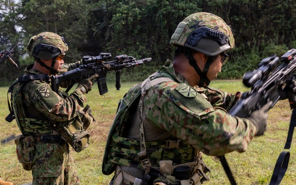 Military personnel in camouflage uniforms are conducting what appears to be a weapons training or demonstration exercise in an outdoor field setting. The soldiers are wearing tactical gear including helmets and body armor, and are handling rifles in various positions. A mix of personnel with different uniform patterns suggests a multinational or joint training exercise.