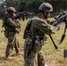 Military personnel in camouflage uniforms are conducting what appears to be a weapons training or demonstration exercise in an outdoor field setting. The soldiers are wearing tactical gear including helmets and body armor, and are handling rifles in various positions. A mix of personnel with different uniform patterns suggests a multinational or joint training exercise.