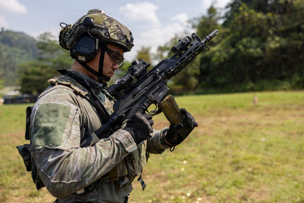 A soldier in full tactical gear is handling a belt-fed light machine gun in an outdoor field setting. The individual is equipped with modern military equipment including a tactical helmet, hearing protection, optical sights, and a plate carrier vest. The setting appears to be a training range or exercise area with green vegetation in the background.