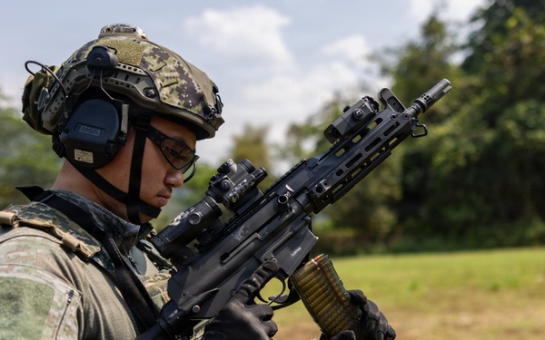 A soldier in full tactical gear is handling a belt-fed light machine gun in an outdoor field setting. The individual is equipped with modern military equipment including a tactical helmet, hearing protection, optical sights, and a plate carrier vest. The setting appears to be a training range or exercise area with green vegetation in the background.