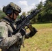 A soldier in full tactical gear is handling a belt-fed light machine gun in an outdoor field setting. The individual is equipped with modern military equipment including a tactical helmet, hearing protection, optical sights, and a plate carrier vest. The setting appears to be a training range or exercise area with green vegetation in the background.