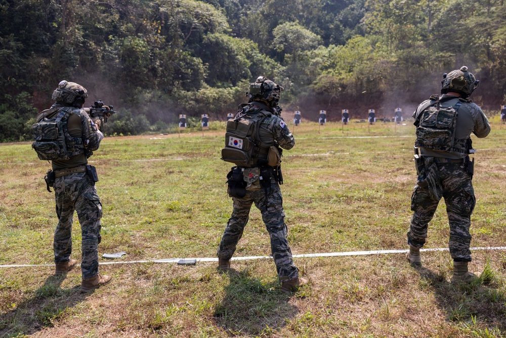 Three military personnel in camouflage uniforms are conducting a live-fire shooting exercise on an outdoor range. At least one soldier is visibly firing a weapon toward a row of human-silhouette targets downrange. The center soldier displays a South Korean flag patch on their backpack, indicating South Korean military affiliation.