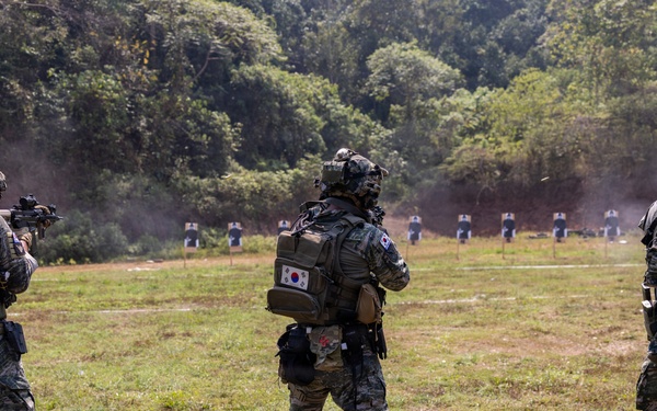 Three military personnel in camouflage uniforms are conducting a live-fire shooting exercise on an outdoor range. At least one soldier is visibly firing a weapon toward a row of human-silhouette targets downrange. The center soldier displays a South Korean flag patch on their backpack, indicating South Korean military affiliation.
