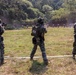 Three military personnel in camouflage uniforms are conducting a live-fire shooting exercise on an outdoor range. At least one soldier is visibly firing a weapon toward a row of human-silhouette targets downrange. The center soldier displays a South Korean flag patch on their backpack, indicating South Korean military affiliation.