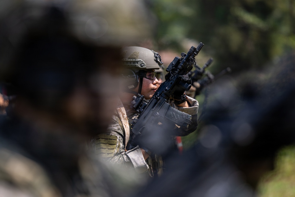 A soldier in full combat gear is visible in sharp focus between blurred foreground and background figures. The individual is wearing a tactical helmet, protective eyewear, body armor, and camouflage uniform while holding a rifle with an optic sight. The shallow depth of field suggests this is a deliberate photographic composition capturing a military formation or exercise.