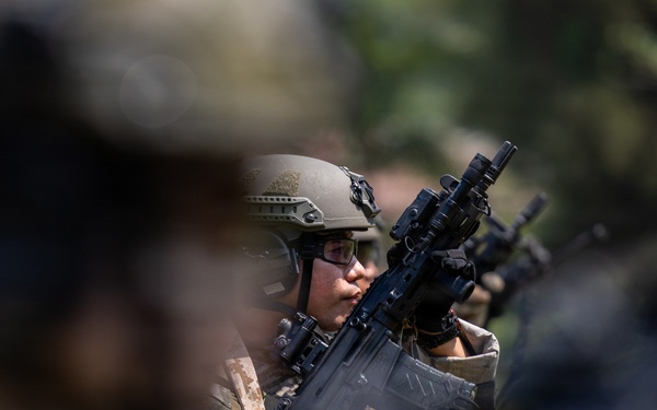 A soldier in full combat gear is visible in sharp focus between blurred foreground and background figures. The individual is wearing a tactical helmet, protective eyewear, body armor, and camouflage uniform while holding a rifle with an optic sight. The shallow depth of field suggests this is a deliberate photographic composition capturing a military formation or exercise.