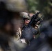 A soldier in full combat gear is visible in sharp focus between blurred foreground and background figures. The individual is wearing a tactical helmet, protective eyewear, body armor, and camouflage uniform while holding a rifle with an optic sight. The shallow depth of field suggests this is a deliberate photographic composition capturing a military formation or exercise.