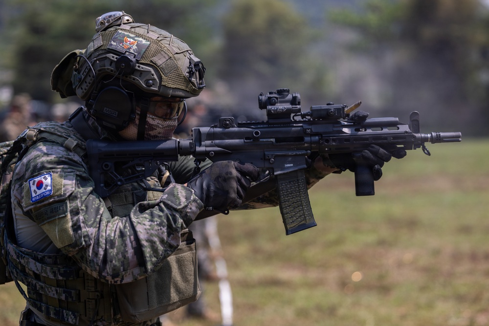 A Republic of Korea (ROK) Army soldier is shown in a combat-ready stance, aiming a rifle with optical sights attached. The soldier is wearing full tactical gear including a camouflage uniform, tactical helmet with accessories, ear protection, and a face mask. The setting appears to be an outdoor training or exercise environment.