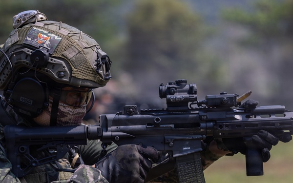A Republic of Korea (ROK) Army soldier is shown in a combat-ready stance, aiming a rifle with optical sights attached. The soldier is wearing full tactical gear including a camouflage uniform, tactical helmet with accessories, ear protection, and a face mask. The setting appears to be an outdoor training or exercise environment.