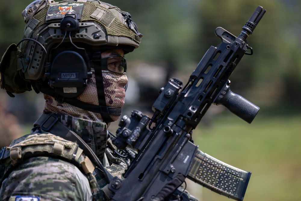 A heavily equipped military or special operations soldier is shown in a close-up profile view, wearing full tactical gear including a camouflage helmet, MSA hearing protection, and a face covering. The soldier is holding a modern assault rifle with visible accessories including an optic sight and what appears to be a loaded magazine. The background is blurred, suggesting an outdoor field or training environment.