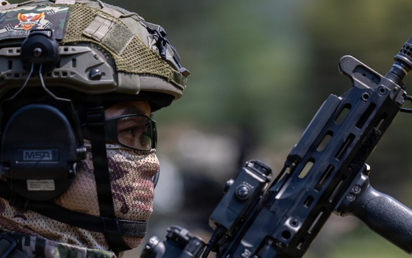 A heavily equipped military or special operations soldier is shown in a close-up profile view, wearing full tactical gear including a camouflage helmet, MSA hearing protection, and a face covering. The soldier is holding a modern assault rifle with visible accessories including an optic sight and what appears to be a loaded magazine. The background is blurred, suggesting an outdoor field or training environment.